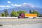 Burgundy long hauler big rig semi truck with orange dry van semi trailer driving on the flat highway road with snow mountain on