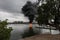 Burning boat with heavy smoke on the water surrounded by trees and buildings under a cloudy sky