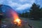 Burning camp fire into remote larch and pine tree woodland with high altitude landscape and dramatic sky at dusk. Summer adventure