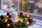 Burning white candle in fir wreath decorated with red Christmas balls and coiled with glowing garland with warm light on white kni