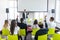 Business coach. Young man gesturing with hand while standing against defocused group of people sitting at the chairs in front of