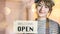 Business owner attractive young Asian woman in apron hanging we`re open sign on front door smiling welcoming clients to new cafe.