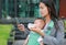 Business woman using telephone with carrying her infant and feeding her child`s by milk bottle. Busy mother hurrying in the