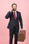 Businessman in black suit and red tie with vintage briefcase talking over phone on pink wall background.