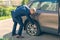 A businessman in a blue suit crouching near his car and checks the degree of damage to a punctured wheel. Hole in the tire. Concep