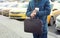 Businessman in casual jacket with leather bag and cup of coffee is standing on city road with taxi in the background and preparing