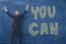 Businessman happily standing with his arms up near the words `you can` written on the dark blue wall.