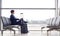 Businessman With Luggage Sitting In Airport Departure Lounge Looking Out Of Window