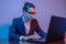 Businessman in a medical mask works at a laptop in the workplace in neon light. Crisis for business during the pandemic