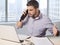 Businessman at office in front of skyscraper window view talking upset on the phone working with computer