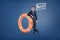 A businessman stands leaning on a large orange life buoy with a speech bubble holding a word Help inside.