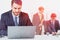 Businessman in a suit working at office desk in front of laptop while two other collegues behind are discussing project. Concept