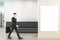 Businessman walking in modern concrete office interior with wooden flooring, black reception desk and white mock up poster on wall