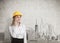 Businesswoman wearing a yellow hard hat and talking on her smartphone while standing near a concrete wall with a city panorama.