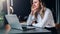 Businesswoman in white shirt is sitting in office at table in front of computer and pensively looks at screen of laptop