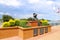 A bust statue of a Spanish explorer on the Savannah riverfront surrounded by red brick and lush green plants and yellow flowers