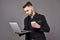 Busy bearded man in business clothes talking by smartphone and using laptop computer while holding cup of coffee in hand