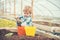 Busy kid filling orange and yellow pots with soil. Blond boy playing with spade in greenhouse