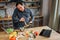 Busy man stand at table in kitchen and blend eggs. He talk on phone and look down. Colorful vegetables lying on desk.