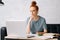 Busy pretty redhead young woman student is taking notes in workbook by looking at laptop screen.