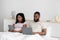 Busy serious millennial black wife and husband working on laptop and tablet, sitting on bed