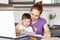 Busy working mother sits in front of opened laptop computer, tries to conecntrate on work, sits against kitchen interior with chil