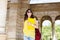 Buttom view pretty brunette girl in sunglasses walking with yellow laptop on archway background. She wears vinous bag