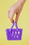 Buying things at market shops concept. Woman hand holding small tiny shopping basket trolley over trend yellow background