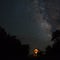 Cabin with it`s windows glowing bright under the milky way at night and surrounded by the silhouetted oak trees