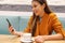 Cafe city lifestyle girl with phone drinking cappuccino reading message on smartphone app sitting indoor in trendy urban cafe