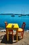 Cafe tableon beach in Adamantas town on Milos island with Aegean sea with boats in background