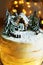 Cake with white cream with gingerbread house and fir trees on a background of Christmas lights