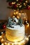Cake with white cream with gingerbread house and fir trees on a background of Christmas lights