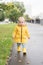 Calm funny toddler baby girl 2 years old in yellow raincoat walking on the asphalt road in raining day. Stylish girl is wearing