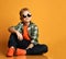 Calm nice kid, schoolboy teenager in round sunglasses, checkered shirt, t-shirt and jeans sits on floor looking up