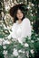 Calm portrait of beautiful hipster girl standing in white blooms in spring. Stylish boho woman in hat posing in blooming tree with