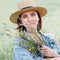 Calm portrait of a young woman with wildflowers bouquet. She dressed a jeans jacket, straw hat and light summer dress. Natural