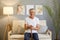 Calm smiling grey-haired mature man wearing casual white T-shirt sitting on sofa at home interior holding his wooden cheek resting