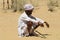 Camel herder wearing wight clothes sits on the sand in Thar desert in Jamba, India.