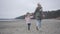 Camera following adult woman and little girl in warm clothes running on the autumn beach. Joyful Caucasian mother and
