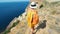 Camera following woman in yellow dress and hat walking to cliff edge over seashore