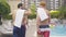 Camera follows confident athletic men walking along the pool at luxurious resort. Back view of young male friends of