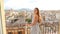 Camera follows young woman welcoming with summer dress and waving long hair from apartment to balcony with beautiful cityscape of 