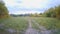 Camera moves above a forest dirt road among dense trees with green foliage in and then he drives out to the autumn meadow