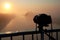 A camera on a tripod stands on a bridge over a river during a sunrise