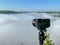 A camera with a tripod at sunrise looks at a foggy canyon.