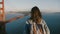 Camera zooms in behind young happy woman with backpack, hair in the wind enjoying sunset scenery at Golden Gate Bridge.