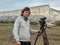 Cameraman with camera on the tripod with long dark hair in a grey jacket smiling against of white cliffs of Mount Ak-Kaya.