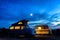 A campervan and a caravan illuminated from inside at nightfall with the moon glowing in a stormy sky.