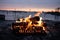 Campfire with bright orange flames blazing on gray rocks near a body of blue water